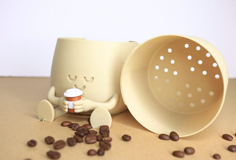 May include: A beige ceramic coffee cup holder with a smiling face and two arms holding a small brown coffee cup. The holder has a removable beige lid with holes for drainage. The holder is sitting on a brown surface with coffee beans scattered around it.