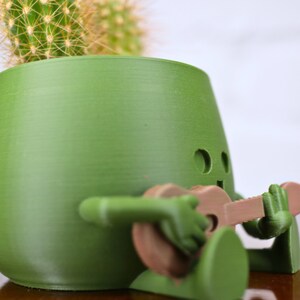 May include: A green ceramic planter shaped like a person playing a guitar. The planter has a cactus growing inside.