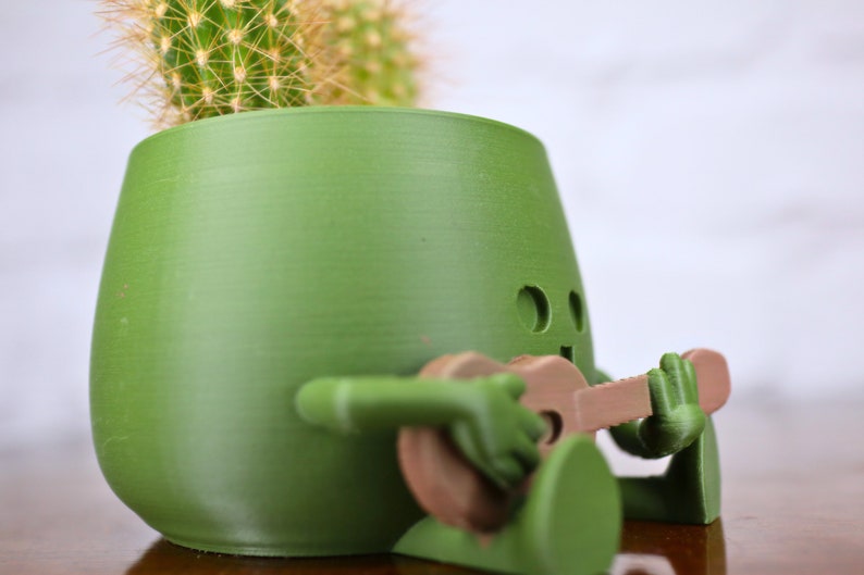 May include: A green ceramic planter shaped like a person playing a guitar. The planter has a cactus growing inside.