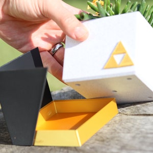 May include: A white planter with a gold triangular symbol, resembling the Triforce from the Legend of Zelda video game series, is being lifted from a black and yellow box. The planter contains a small succulent plant.
