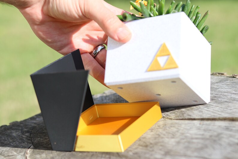 May include: A white planter with a gold triangular symbol, resembling the Triforce from the Legend of Zelda video game series, is being lifted from a black and yellow box. The planter contains a small succulent plant.
