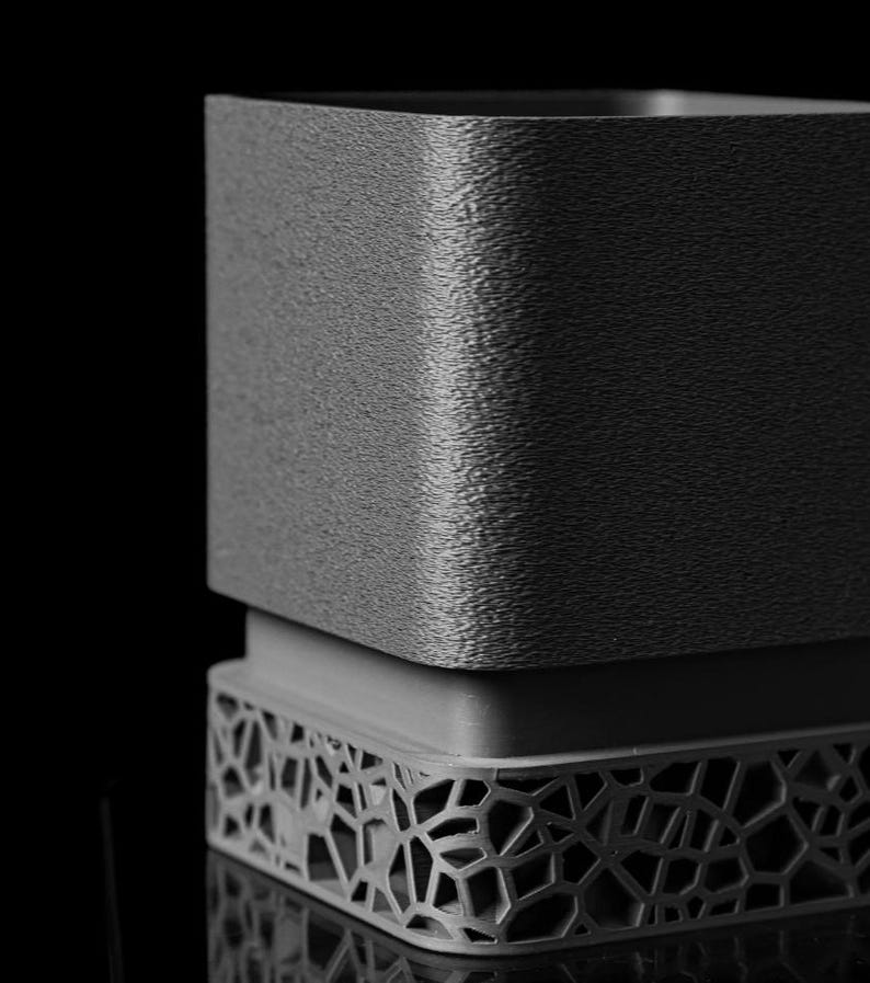 May include: A grayscale, textured, square-shaped container with a decorative, openwork base. The top portion has a rough, grainy surface, while the base features a geometric, web-like pattern. The object is set against a black background.