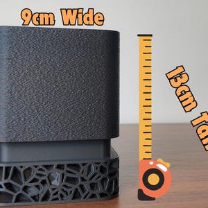 May include: A dark gray, rectangular object with a textured surface, 3.5 inches tall and 3.5 inches wide. The base has a decorative, geometric pattern. A yellow and orange tape measure is next to the object.