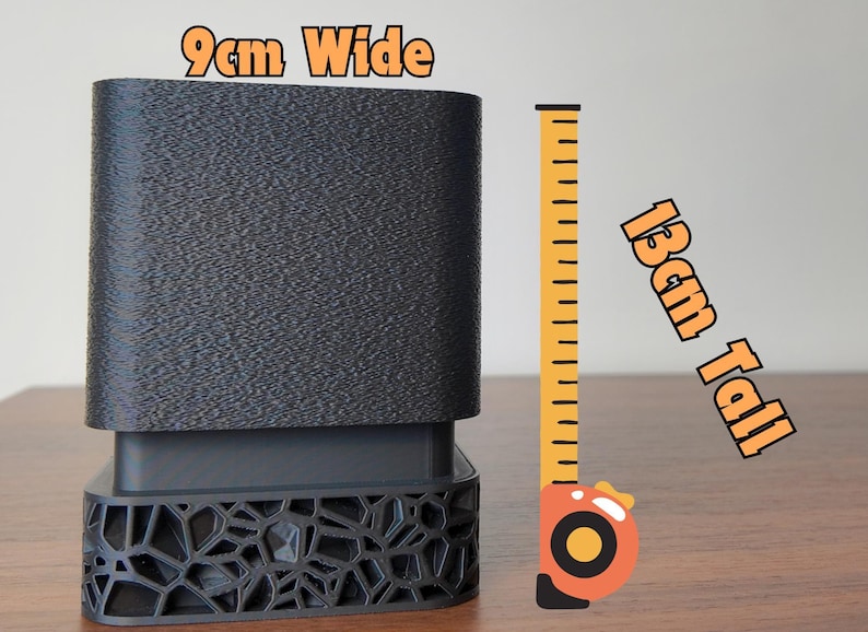 May include: A dark gray, rectangular object with a textured surface, 3.5 inches tall and 3.5 inches wide. The base has a decorative, geometric pattern. A yellow and orange tape measure is next to the object.