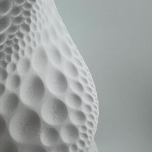 May include: A close-up of a white, textured object with a pattern of rounded, bubble-like shapes. The object has a smooth, curved surface and is set against a light gray background. The design creates a sense of depth and dimension.