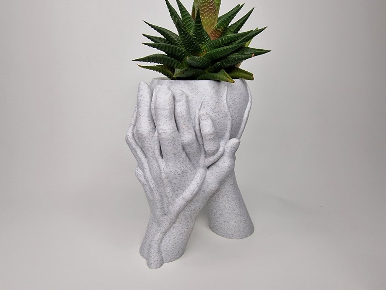 Puede incluir: Maceta de cer&aacute;mica blanca con forma de dos manos sosteniendo una planta suculenta. La maceta tiene una superficie texturizada.