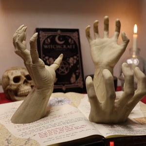 May include: Three skeletal hands reaching up from an open book with the title "Witchcraft" on the cover. The book is on a red surface with two lit candles and a skull in the background.