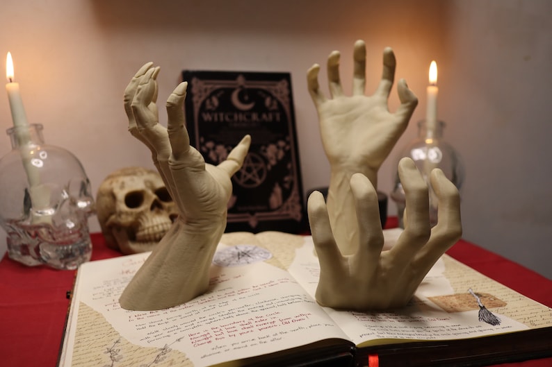 May include: Three skeletal hands reaching up from an open book with the title "Witchcraft" on the cover. The book is on a red surface with two lit candles and a skull in the background.