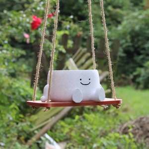 May include: A white ceramic planter shaped like a smiling face hangs from a rope swing. The planter has small feet and is sitting on a red wooden swing seat.