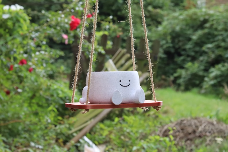 May include: A white ceramic planter shaped like a smiling face hangs from a rope swing. The planter has small feet and is sitting on a red wooden swing seat.