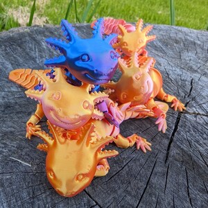 May include: Four 3D printed axolotl figurines, one blue, one orange, and two pink, are stacked on top of each other on a wooden surface.
