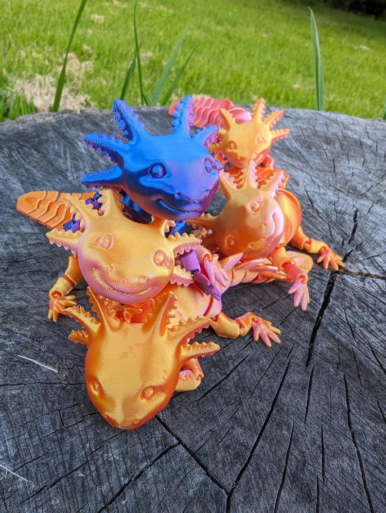 May include: Four 3D printed axolotl figurines, one blue, one orange, and two pink, are stacked on top of each other on a wooden surface.