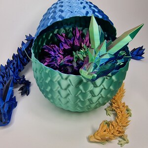 May include: Three origami dragons, one blue, one yellow, and one green, with iridescent scales. The green dragon is inside a teal dragon egg with a blue top.