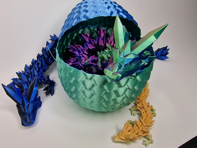 May include: Three origami dragons, one blue, one yellow, and one green, with iridescent scales. The green dragon is inside a teal dragon egg with a blue top.