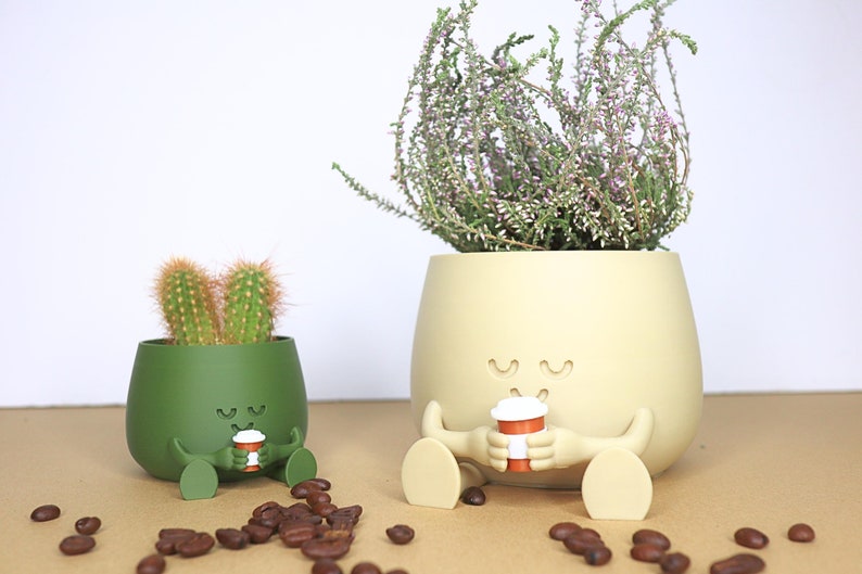 May include: Two green and beige ceramic planters shaped like cartoon characters with closed eyes and smiling mouths. The planters are sitting on a brown surface with coffee beans scattered around them. The planters are holding small coffee cups.