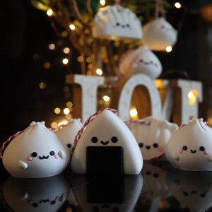 May include: A group of white ceramic ornaments shaped like dumplings and onigiri, with cute faces and pink cheeks. The ornaments are hanging from red and white string and are displayed on a black surface in front of a wooden sign that says "LOVE".