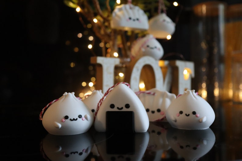 May include: A group of white ceramic ornaments shaped like dumplings and onigiri, with cute faces and pink cheeks. The ornaments are hanging from red and white string and are displayed on a black surface in front of a wooden sign that says "LOVE".