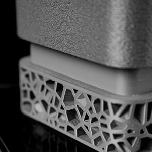 May include: Close-up of a gray, geometric-patterned object. The object has a textured top surface and a detailed, openwork base. The design features a series of interconnected shapes, creating a modern, architectural aesthetic.