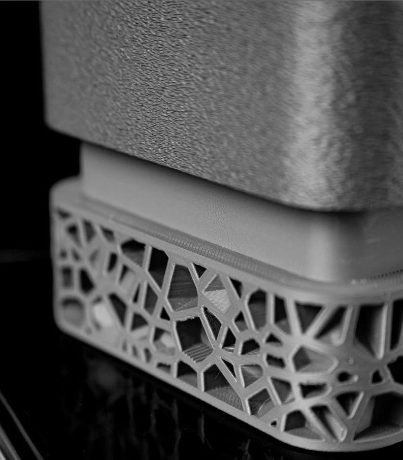 May include: Close-up of a gray, geometric-patterned object. The object has a textured top surface and a detailed, openwork base. The design features a series of interconnected shapes, creating a modern, architectural aesthetic.