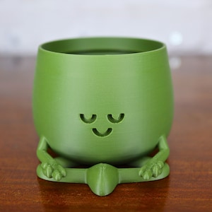 Puede incluir: Un macetero de cer&aacute;mica verde con forma de figura meditando con una cara sonriente. El macetero tiene una abertura ancha en la parte superior para plantar.
