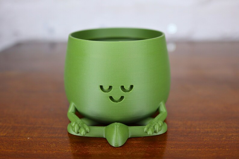 Puede incluir: Un macetero de cer&aacute;mica verde con forma de figura meditando con una cara sonriente. El macetero tiene una abertura ancha en la parte superior para plantar.