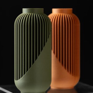 Peut inclure: Deux vases d&eacute;coratifs. L'un est vert olive, l'autre est orange. Les deux ont une forme cylindrique avec des d&eacute;tails verticaux c&ocirc;tel&eacute;s et un &eacute;l&eacute;ment de design diagonal. Les vases sont plac&eacute;s sur un fond noir.