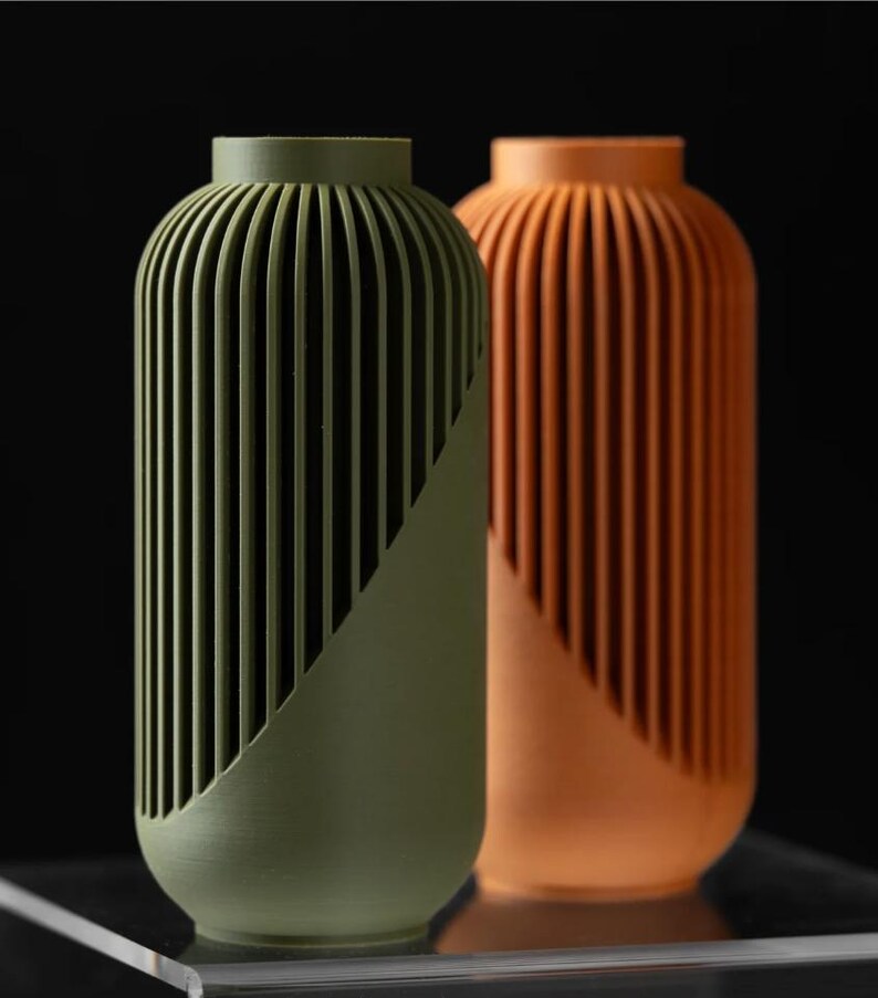 Peut inclure: Deux vases d&eacute;coratifs. L'un est vert olive, l'autre est orange. Les deux ont une forme cylindrique avec des d&eacute;tails verticaux c&ocirc;tel&eacute;s et un &eacute;l&eacute;ment de design diagonal. Les vases sont plac&eacute;s sur un fond noir.