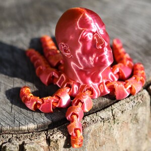 May include: A 3D printed sculpture of a human head with a metallic red finish, mounted on a base of multiple 3D printed tentacles.