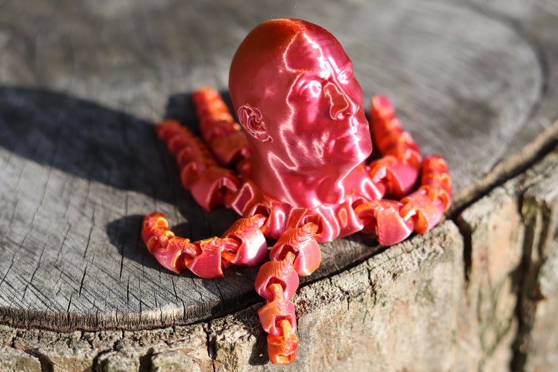 May include: A 3D printed sculpture of a human head with a metallic red finish, mounted on a base of multiple 3D printed tentacles.
