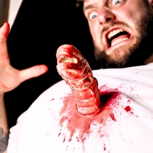 May include: A man with a beard is screaming in fear as a bloody alien creature bursts from his chest. The creature has sharp teeth and a fleshy, segmented body.
