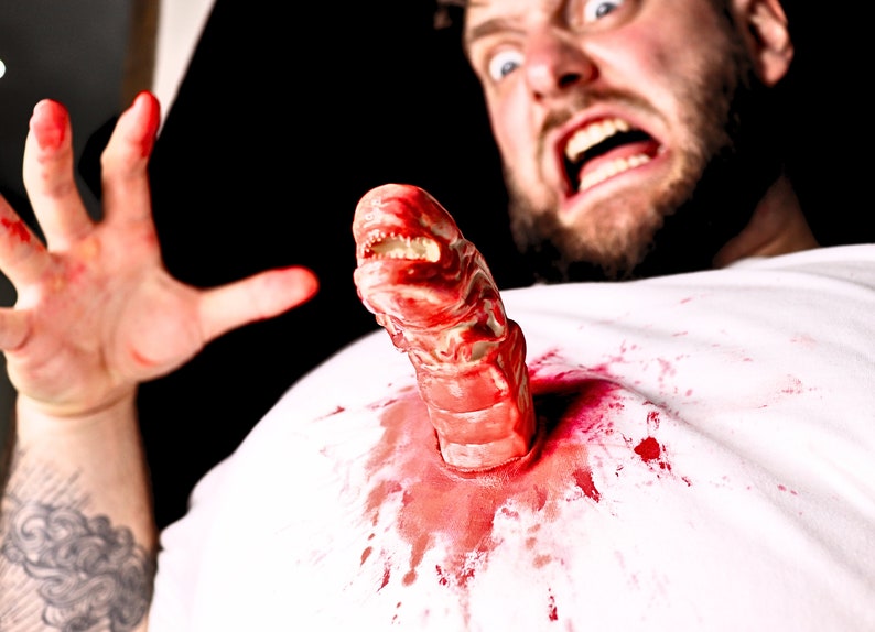 May include: A man with a beard is screaming in fear as a bloody alien creature bursts from his chest. The creature has sharp teeth and a fleshy, segmented body.