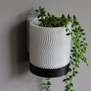 May include: White wall-mounted planter with a wavy texture and a black base. A trailing green plant is growing in the planter.