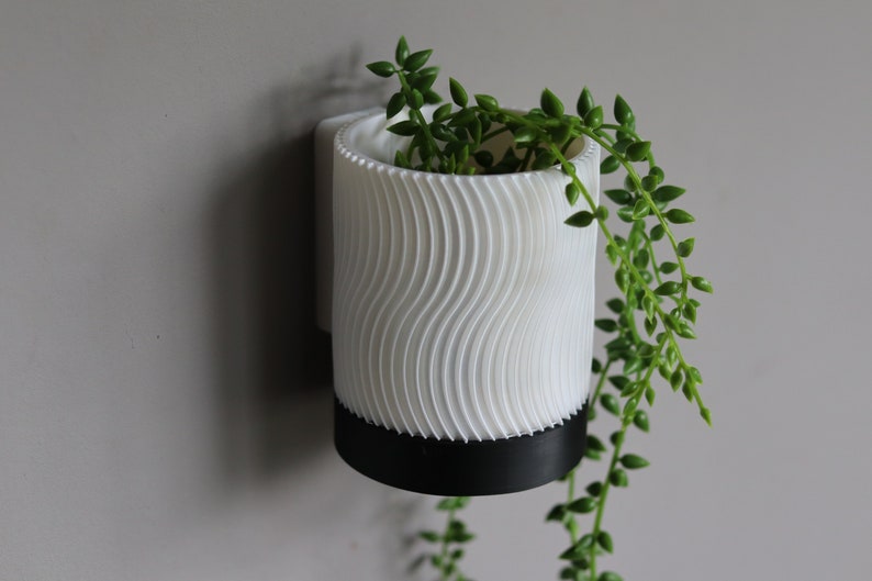 May include: White wall-mounted planter with a wavy texture and a black base. A trailing green plant is growing in the planter.