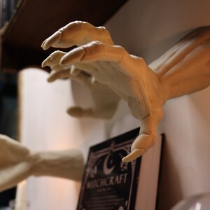 May include: Two white plaster hands with long fingers are mounted on a wall. The hands are reaching out, as if to grab something. A book with the title "Witchcraft" is visible in the background.