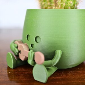 May include: A green 3D printed planter shaped like a person playing a guitar. The planter has a small cactus growing inside.