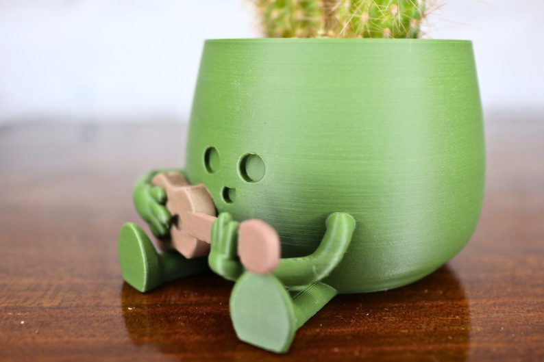 May include: A green 3D printed planter shaped like a person playing a guitar. The planter has a small cactus growing inside.