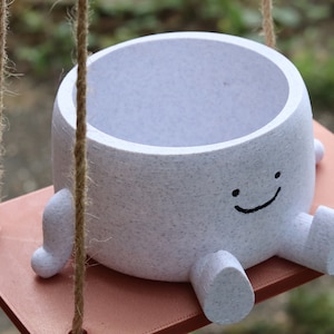 May include: White ceramic planter with a smiling face and small legs, hanging from a rope swing. The planter is on a light brown wooden platform.