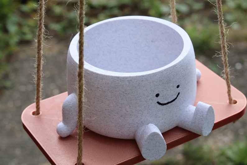 May include: White ceramic planter with a smiling face and small legs, hanging from a rope swing. The planter is on a light brown wooden platform.