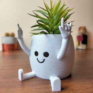 Peut inclure: Un pot en céramique blanc en forme de personne souriante avec les bras et les jambes tendus. Le pot contient une petite plante verte.