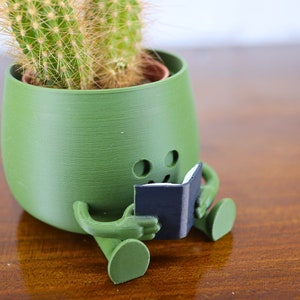 Puede incluir: Un macetero de cer&aacute;mica verde con forma de persona sentada con las piernas cruzadas y sosteniendo un peque&ntilde;o libro negro. El macetero tiene una planta de cactus en su interior.