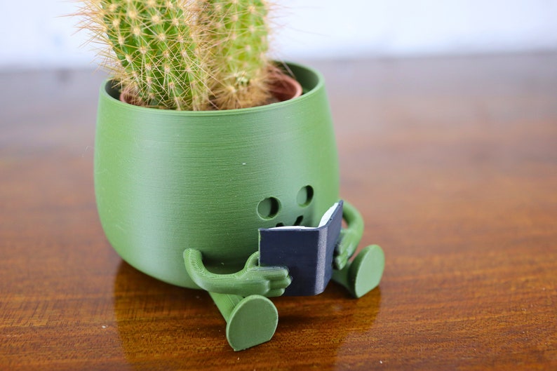 Puede incluir: Un macetero de cer&aacute;mica verde con forma de persona sentada con las piernas cruzadas y sosteniendo un peque&ntilde;o libro negro. El macetero tiene una planta de cactus en su interior.