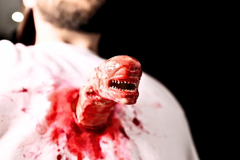 May include: A close-up of a bloody, fleshy alien creature bursting out of a person's chest. The creature has a large mouth with sharp teeth and is partially visible.