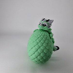 Puede incluir: Una figurita de drag&oacute;n verde con detalles blancos y negros, enroscada alrededor de un huevo verde con textura escamosa.
