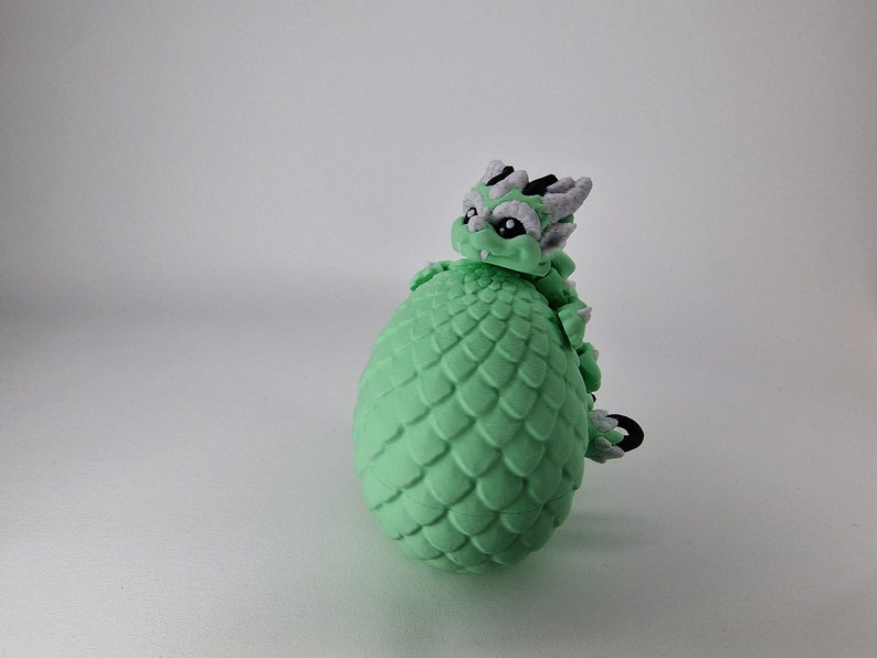 Puede incluir: Una figurita de drag&oacute;n verde con detalles blancos y negros, enroscada alrededor de un huevo verde con textura escamosa.