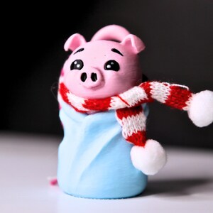 May include: A pink pig figurine wearing a red and white striped scarf with white pom-poms. The pig is wrapped in a blue blanket.