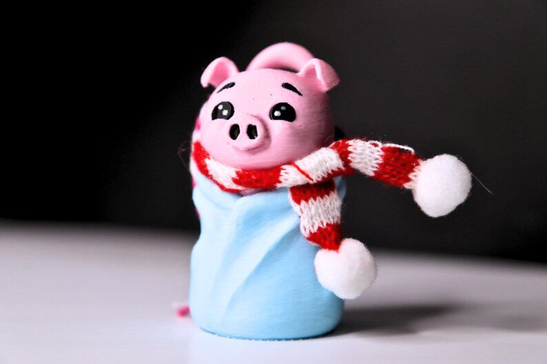 May include: A pink pig figurine wearing a red and white striped scarf with white pom-poms. The pig is wrapped in a blue blanket.