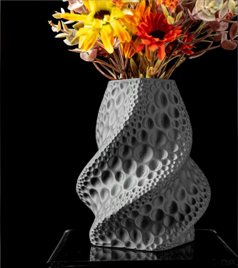 May include: A gray, textured vase with a spiral design, filled with colorful artificial flowers. The vase has a unique pattern of raised circles and a twisted shape. The flowers include yellow, orange, and red blooms.
