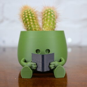 Puede incluir: Un macetero de cer&aacute;mica verde con forma de persona sentada con las piernas cruzadas, sosteniendo un libro negro. Dos peque&ntilde;os cactus crecen en la parte superior del macetero, pareciendo orejas.
