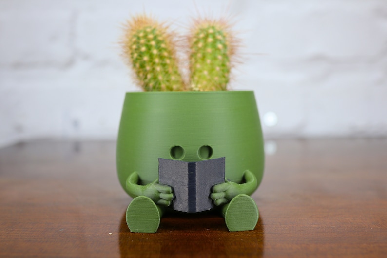 Puede incluir: Un macetero de cer&aacute;mica verde con forma de persona sentada con las piernas cruzadas, sosteniendo un libro negro. Dos peque&ntilde;os cactus crecen en la parte superior del macetero, pareciendo orejas.