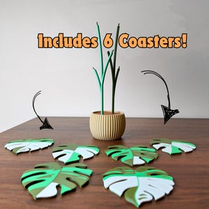 May include: A set of six leaf-shaped coasters in shades of green and white, arranged on a wooden surface. A decorative vase with green stems is in the background. The text "Includes 6 Coasters!" is displayed in orange.